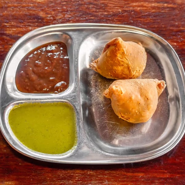 Two golden-brown samosas and two chutneys, green and red, on a metal plate.